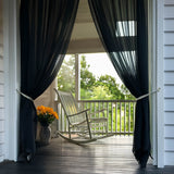 Black sheer curtains drape across an outdoor porch that overlooks at rocking chair.