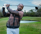 A man swings a golf club on a beautiful green golf course with a pond behind. He's wearing a sheer black mosquito net jacket.