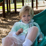 A blond child smiles while he sits at the bottom of a slide. He's dressed in a ShrugBug mosquito net suit.