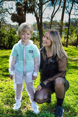 A little blond boy stands on bright green grass with a young adult female crouching beside him. They are both wearing full mosquito net gear to protect from insects. He is wearing a white suit, she is wearing a black suit.